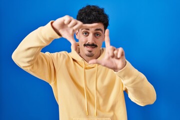 Fototapeta premium Hispanic man standing over blue background smiling making frame with hands and fingers with happy face. creativity and photography concept.