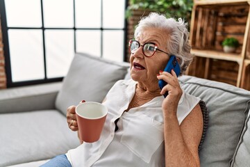 Senior grey-haired woman drinking coffee and talking on the smartphone sitting on sofa at home