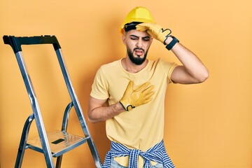 Handsome man with beard by construction stairs wearing hardhat touching forehead for illness and fever, flu and cold, virus sick