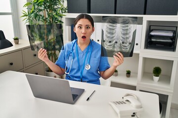 Young latin doctor woman holding x-ray afraid and shocked with surprise and amazed expression, fear and excited face.