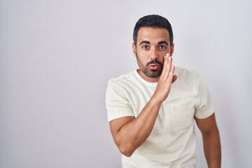 Hispanic man with beard standing over isolated background hand on mouth telling secret rumor, whispering malicious talk conversation