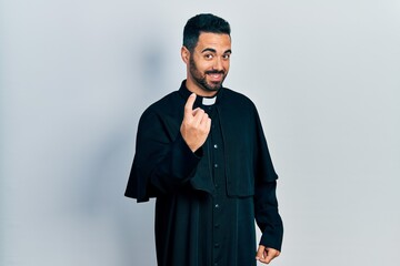 Handsome hispanic man with beard wearing catholic priest robe beckoning come here gesture with hand...