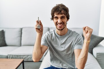 Young hispanic man holding keys of new home sitting on the sofa screaming proud, celebrating victory and success very excited with raised arm