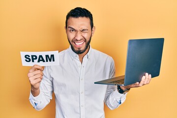 Hispanic man with beard working using computer laptop holding spam banner winking looking at the camera with sexy expression, cheerful and happy face.