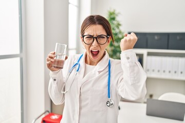 Young brunette doctor woman holding glass of water annoyed and frustrated shouting with anger,...