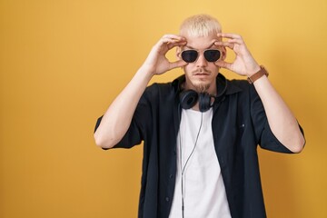 Young caucasian man wearing sunglasses standing over yellow background trying to open eyes with fingers, sleepy and tired for morning fatigue