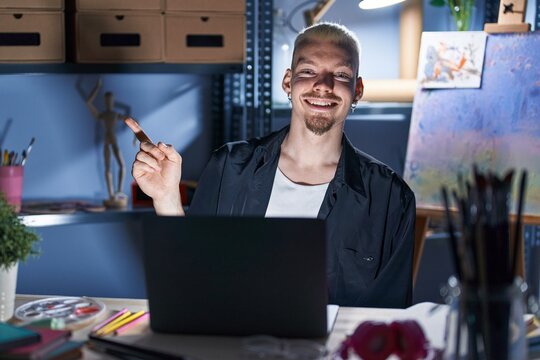 Young caucasian man using laptop at night at art studio with a big smile on face, pointing with hand and finger to the side looking at the camera.