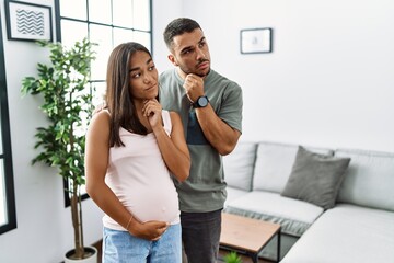 Young interracial couple expecting a baby, touching pregnant belly with hand on chin thinking about...