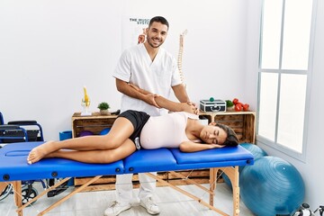 Latin man and woman wearing physiotherapist uniform having pregnancy rehab session stretching arm at physiotherapy clinic