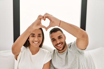 Young hispanic couple doing heart symbol with hands sitting on the bed at home.