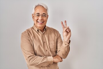 Hispanic senior man wearing glasses smiling with happy face winking at the camera doing victory...