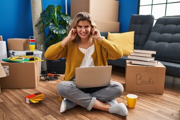 Young woman sitting on the floor at new home using laptop covering ears with fingers with annoyed...