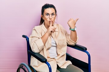 Young hispanic woman sitting on wheelchair asking to be quiet with finger on lips pointing with hand to the side. silence and secret concept.