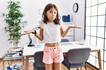 Young hispanic girl standing at pediatrician clinic clueless and confused expression with arms and hands raised. doubt concept.
