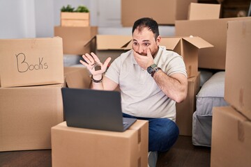 Plus size hispanic man with beard moving to a new home doing video call covering mouth with hand, shocked and afraid for mistake. surprised expression