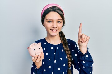 Young brunette girl holding piggy bank smiling with an idea or question pointing finger with happy...