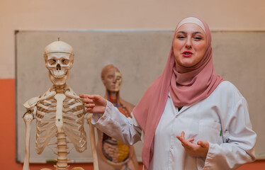 A Muslim female teacher teaches students using the human body skeleton model to explain anatomy in biology school class in the classroom lab.	
