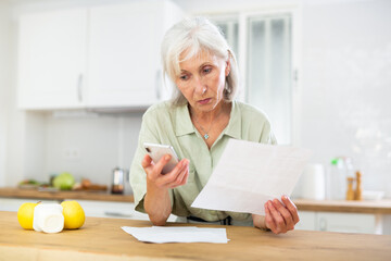 Thoughtful senior woman analyzing bills and using smartphone, standing at kitchen table at home.