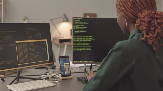 Slowmo portrait of African American female programmer typing program code sitting in front of two monitors at office desk, then turning on chair and smiling at camera