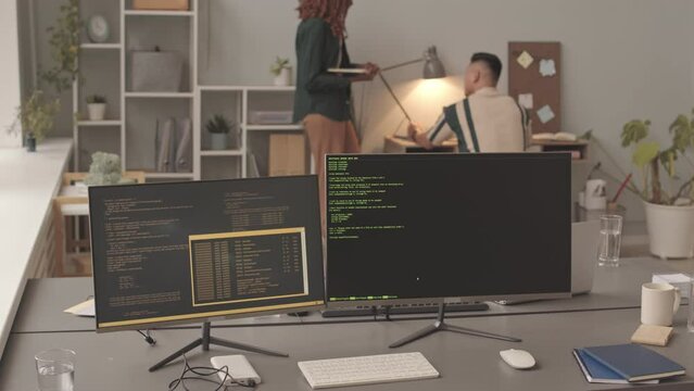 Slowmo of two computer monitors with program codes on screen on table in foreground while diverse couple of programmers working together in background, developing new software