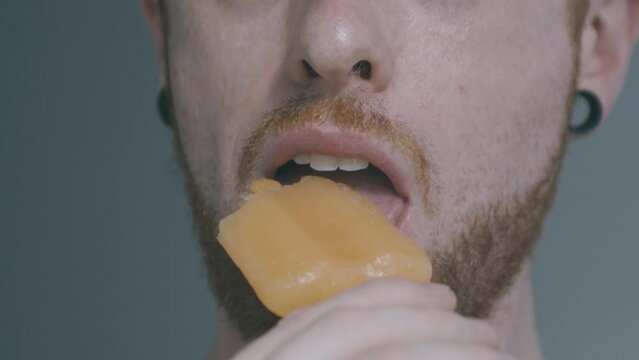 A boy with ear dilators stacks the camera with a peach popsicle, and then approaches the popsicle and eats it
