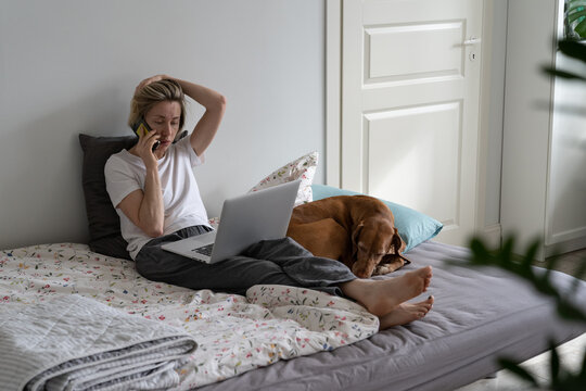 Busy Middle-aged Woman Talks On Smartphone With Important Person About Job Vacancy. Blonde Female Looks Concerned And Anxious Talking With Potential Employer Sitting On Bed Near Sleeping Dog