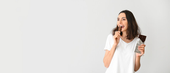 Beautiful young woman eating tasty chocolate on light background with space for text