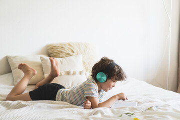 Cute boy lying on the bed at home and playing with pc tablet or reading online and listening to...