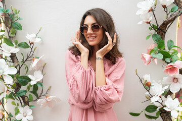Obraz na płótnie Canvas Young beautiful smiling cute romantic woman in trendy summer pink dress. Carefree woman posing in the street near white wall with flowers. Positive model outdoors in sunglasses.