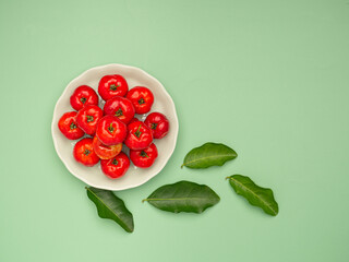 Top view of ripe red acerola cherries on a white dish and green leaves over a green background
