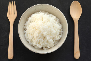 White rice in bowl on a black wooden background. Steam rice in bowl and wooden spoon, fork on the table.Top view 