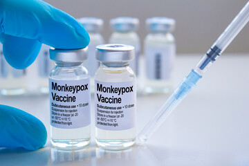 A healthcare worker holding a Monkeypox vaccine vial next to a syringe on a white table. 