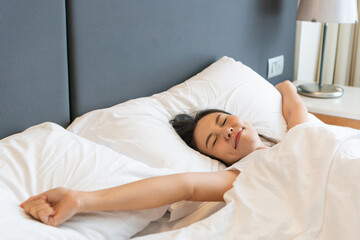 Fototapeta na wymiar Smiling young Asian woman stretching after wake up with sunlight in the morning in bedroom at home or hotel. Closeup, copy space