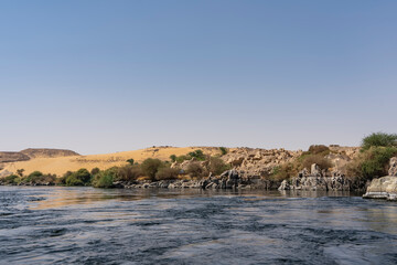 Picturesque boulders and green vegetation are visible along the riverbank. A sand dune against a clear blue sky. Copy space. Egypt. Nile