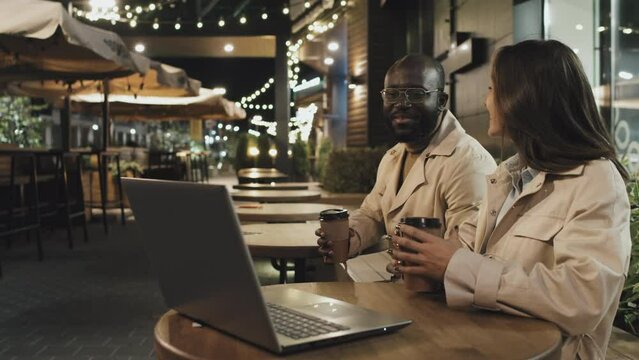 African American man and Caucasian woman sitting at table outdoors late in evening having online meeting with foreign business partners
