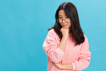 Sad cute Asian student young lady in pink hoodie sweatshirt wear round glasses recline on hand posing isolated on blue studio background. The best offer for ad. Eyewear for vision correction concept
