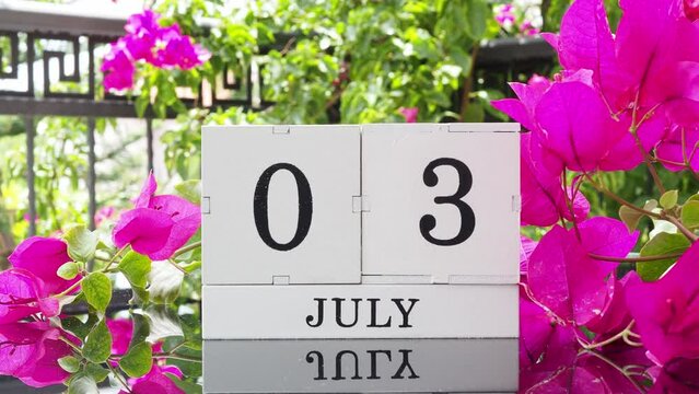 A wooden calendar with an important event for July 3 on a table with hibiscus flowers, a woman's hand sets the date on the calendar. Flip the calendar over to a cubic perpetual calendar. summer month