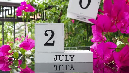 A wooden calendar with an important event for July 20 on a table with hibiscus flowers, a woman's hand sets the date on the calendar. Flip the calendar over to a cubic perpetual calendar. summer month