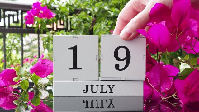 A wooden calendar with an important event for July 19 on a table with hibiscus flowers, a woman's hand sets the date on the calendar. Flip the calendar over to a cubic perpetual calendar. summer month