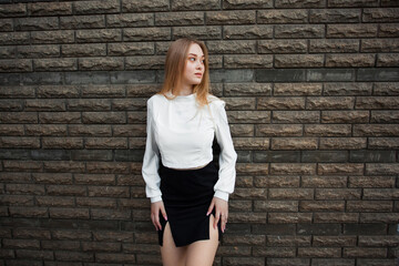 A slender young schoolgirl with blond hair, in a white shirt and a short black skirt, stands against a dark textured stone wall, looks away and keeps her hands on her hips.