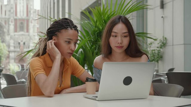 Asian and mixed race girls worried about the news on lap top. Young female students sit outdoors in a summer cafe and prepare for exams