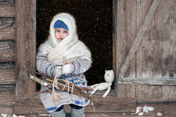 Rustic winter motif. Vintage. A little girl who has returned from the forest is sitting on the threshold of an old village house with a bundle of brushwood on her lap. Blizzard. Copy space.