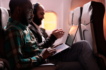 African american male freelancer working on laptop during sunset flight, waiting to arrive at...
