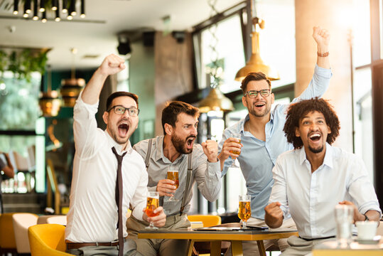 Four young men in casual clothes are cheering for football and holding glasses of beer while sitting in pub