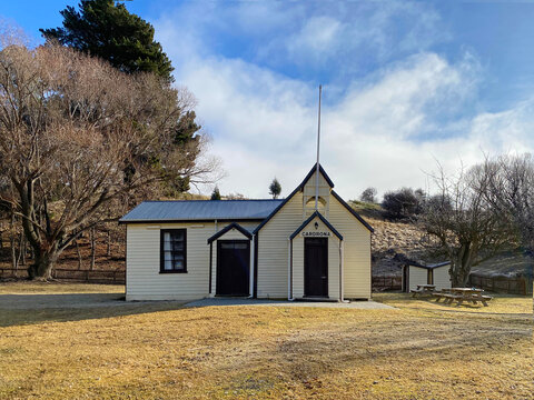 Cardrona village town hall with grass lawn and a picket fence viewed through trees.
