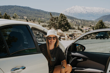 Woman traveler in hat sitting in car with mountain background. Road trip. Young adult woman in sun glasses at summer holiday vacation.