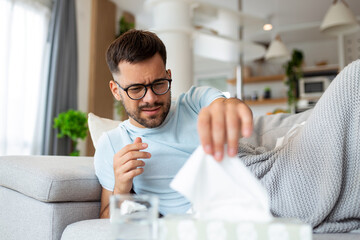 Sick sad man with glasses sits on couch at home suffers from runny nose flu disease coronavirus pandemic covid epidemic sneezes. Unwell guy feeling bad fever virus illness symptoms indoor