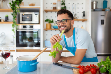 Handsome smiling young man leaning on kitchen counter with vegetables and looking away. Portrait of happy casual guy in apron leaning on steel counter in the kitchen with ingredients on it.