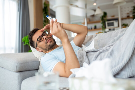 Man feeling sick lying in the bed and looking the thermometer. Sick man lying on sofa checking his temperature under a blanket at home in the living room