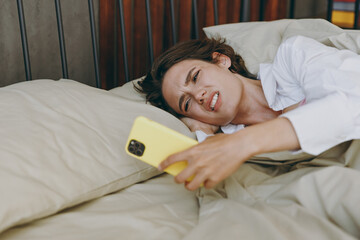 Young sad tired sleepy unhappy woman wear white shirt pajama she lying in bed hold mobile cell phone alarm rest relax spend time in bedroom lounge home in own room hotel wake up Real estate concept.